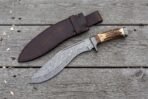 Overhead view of the Damascus kukri with stag horn handle and steel butt cap beside its leather sheath on boards