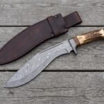 Overhead view of the Damascus kukri with stag horn handle and steel butt cap beside its leather sheath on boards