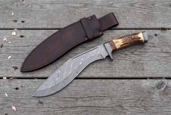 Overhead view of the Damascus kukri with stag horn handle and steel butt cap beside its leather sheath on boards