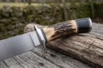 Bowie knife with stag horn and black micarta handle, close view of the steel guard and polished blade resting on a weathered log
