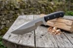 Bushcraft tracker knife with black handle laid on a round wooden table next to split firewood and shavings