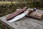 Hunting bowie knife with stag horn handle and steel guard set on an old plank table next to its brown leather sheath