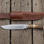 Steel camp knife with stag horn handle and steel guard laid on wooden boards below its brown leather sheath, oak leaves around