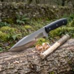 Tracker knife with black micarta handle propped on a mossy log beside a whittled stick and curled wood shavings
