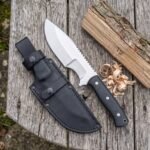 Tracker knife with black micarta handle and saw back spine lying on a wooden table beside its black sheath and wood shavings