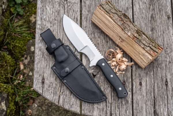 Tracker knife with black micarta handle and saw back spine lying on a wooden table beside its black sheath and wood shavings