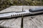 Close view of the tracker knife spine showing the saw back teeth along the polished blade on a wooden table