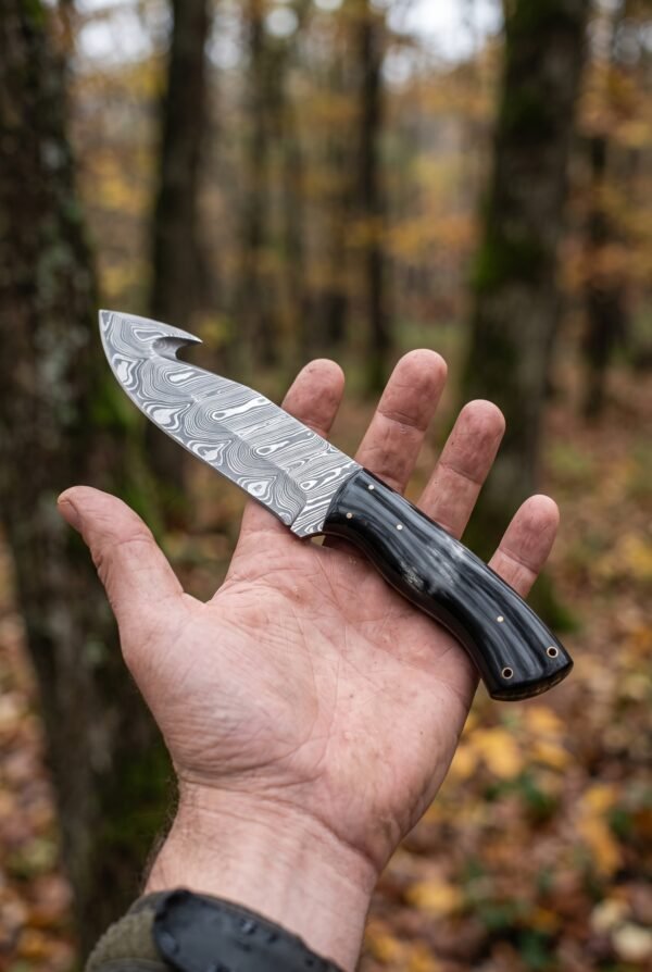 Damascus gut hook hunting knife with black buffalo horn handle held flat on an open palm in an autumn forest