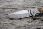 Damascus steel folding knife blade in close view on weathered wood, showing the pattern and the engraved steel bolster
