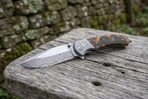 Damascus steel folding knife with resin handle lying open on an old wooden bench beside a mossy stone wall