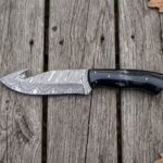 Top view of a Damascus gut hook hunting knife with black horn handle on wooden boards scattered with oak leaves