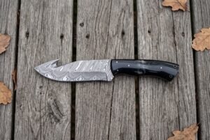 Top view of a Damascus gut hook hunting knife with black horn handle on wooden boards scattered with oak leaves