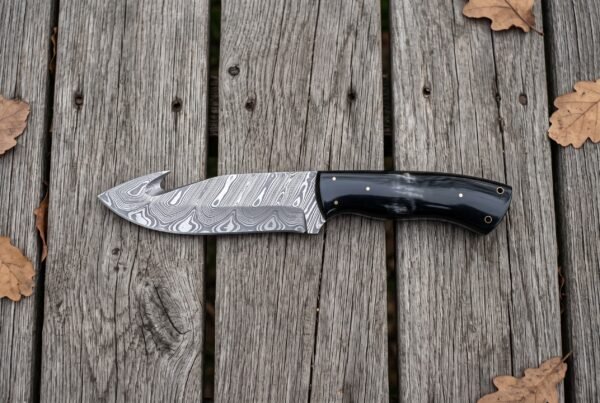 Top view of a Damascus gut hook hunting knife with black horn handle on wooden boards scattered with oak leaves