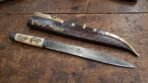 High carbon steel seax lying beside its tooled leather sheath with brass chape and raven mounts on a wooden table