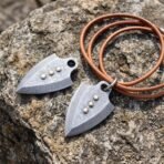 Close view of two Damascus arrowhead pendants, one on a tan leather cord with a steel ring, on grey stone