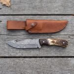 Damascus gut hook knife with stag horn and wenge wood handle seen from above on grey planks with its tan leather sheath