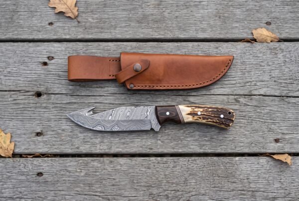 Damascus gut hook knife with stag horn and wenge wood handle seen from above on grey planks with its tan leather sheath