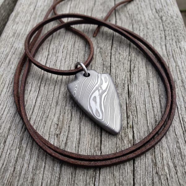 Damascus steel arrowhead pendant on a brown leather cord, laid in loose loops across weathered grey wood