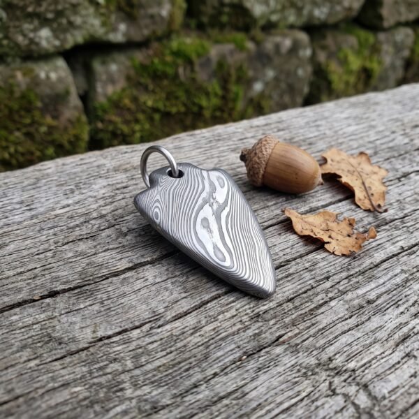 Damascus steel arrowhead pendant on an old wooden log beside an acorn and dry oak leaves, mossy stone wall behind