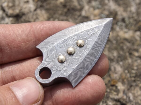 Damascus steel arrowhead pendant with three domed studs held between fingers showing the layered pattern up close
