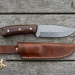 Damascus steel camp knife with pakka wood handle laid on grey boards above its stitched brown leather sheath and shavings