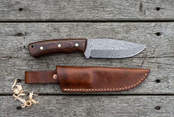 Damascus steel camp knife with pakka wood handle laid on grey boards above its stitched brown leather sheath and shavings