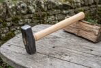 Forged hammer with dark steel head and pale ash wood handle lying on a round wooden table beside a split log