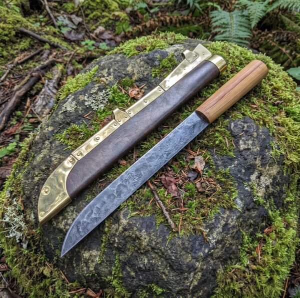 Forged seax knife with light wood handle lying on a mossy boulder next to its brown leather sheath with brass edge fittings