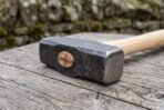 Forged steel hammer head with copper wedge in the eye and ash wood handle, close view on a grey wooden table