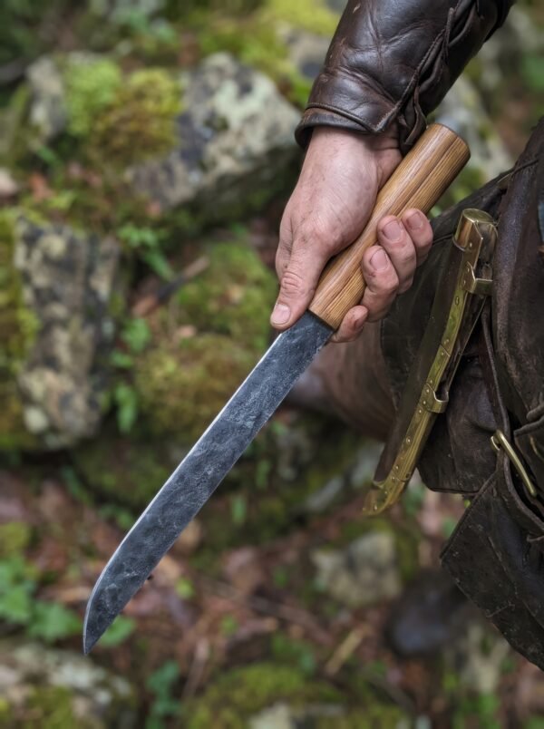 Hand forged carbon steel seax with light wood handle held point down beside a brass fitted leather sheath in a mossy forest