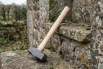 Hand forged hammer with ash wood handle leaning head down against an old mossy stone step outdoors