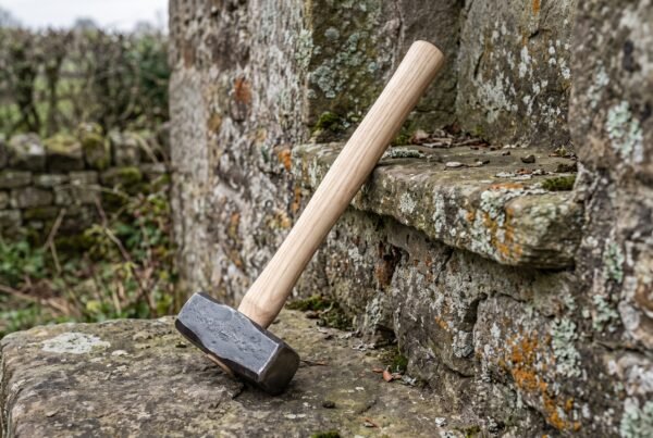 Hand forged hammer with ash wood handle leaning head down against an old mossy stone step outdoors