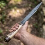 High carbon steel seax held point up with a carved bone handle and copper butt cap against a green forest
