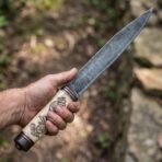 High carbon steel seax knife with carved bone handle showing ravens and runes gripped in hand outdoors near rocks