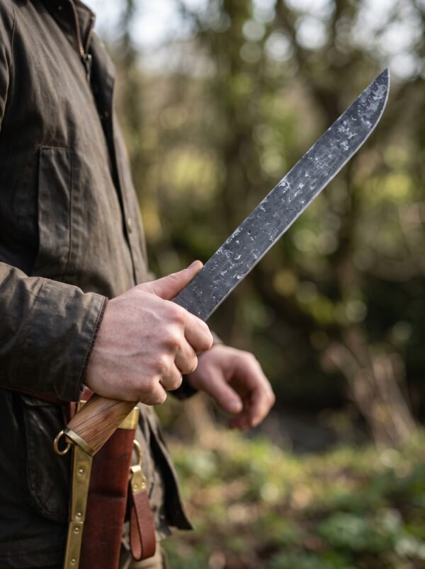 High carbon steel seax knife with wooden handle held in hand outdoors, brass mounted leather sheath worn at the belt