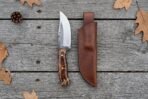High carbon steel skinner knife with pine cone resin handle laid beside its brown leather sheath on grey boards, top view