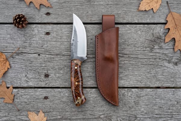 High carbon steel skinner knife with pine cone resin handle laid beside its brown leather sheath on grey boards, top view