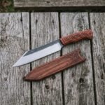 Overhead view of the seax knife and its engraved wooden cover lying side by side on grey wooden boards