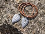 Pair of Damascus steel arrowhead pendants with studs and a tan leather cord laid on a sunlit granite rock