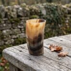 Polished natural horn drinking mug seen at an angle on a weathered wooden rail with fallen leaves and a stone wall