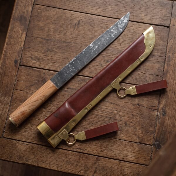 Seax knife with wooden handle lying beside its red leather sheath with brass edging, rings and belt loops on an old table