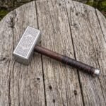 Stainless steel Viking hammer with knotwork engraved head and leather wrapped wooden handle lying on a weathered plank
