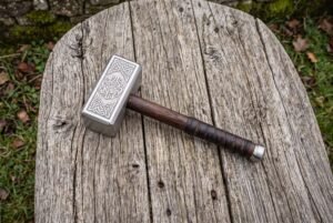 Stainless steel Viking hammer with knotwork engraved head and leather wrapped wooden handle lying on a weathered plank
