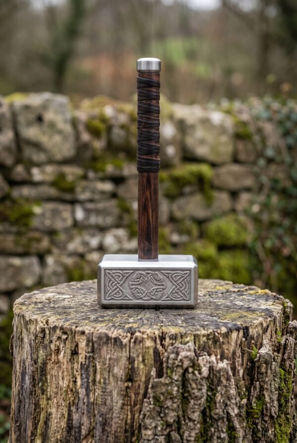 Stainless steel Viking hammer standing upright on its engraved head on a mossy tree stump, leather wrapped handle on top