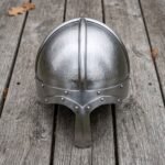Steel Norse helmet with hammered skull and long nasal guard seen from the front on grey wooden decking with fallen leaves