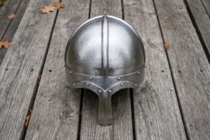 Steel Norse helmet with hammered skull and long nasal guard seen from the front on grey wooden decking with fallen leaves