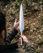 Bright steel Viking spear head held upright point first in both hands above a forest floor, showing the full blade length