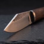 Carbon steel seax blade close up showing rough forge scale along the spine, a black spacer and the wood handle on dark slate