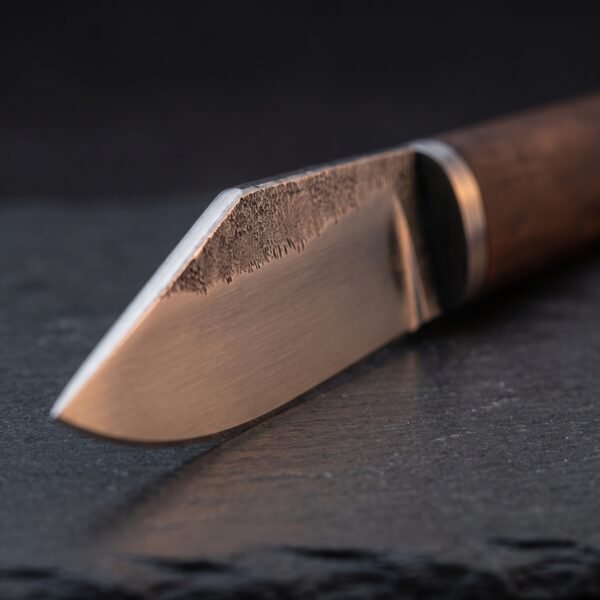Carbon steel seax blade close up showing rough forge scale along the spine, a black spacer and the wood handle on dark slate