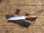 Carbon steel seax knife with a round wood handle lying across its dark brown leather sheath on weathered wooden boards